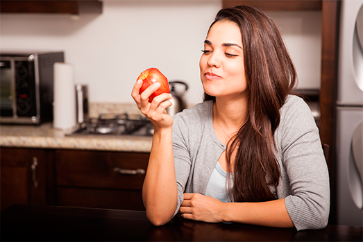 La foto se compone por una mujer joven que come una manzana roja en la barra de la cocina.
